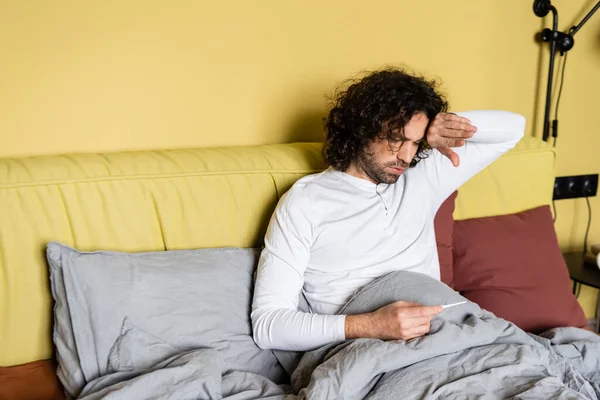 Bouleversé homme touchant le front tout en regardant thermomètre dans le lit — Photo de stock