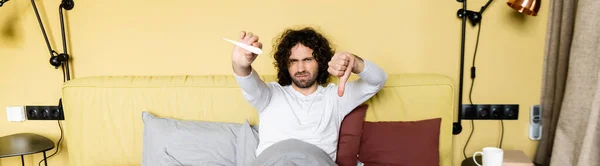 Imagem horizontal do homem descontente segurando o polegar para baixo e olhando para o termômetro na cama — Fotografia de Stock