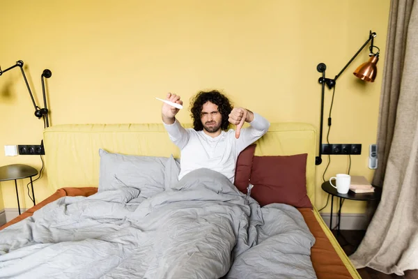 Upset young man showing thumb down while holding thermometer in bed — Stock Photo