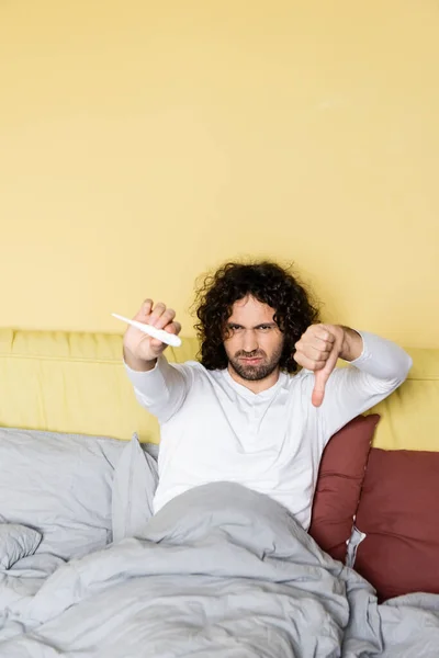 Unzufriedener junger Mann zeigt Daumen nach unten, während er Thermometer im Bett hält — Stockfoto