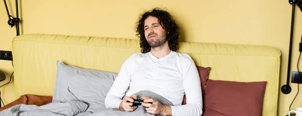KYIV, UKRAINE - APRIL 25, 2020: panoramic crop of dissatisfied young man playing video game in bed — Stock Photo