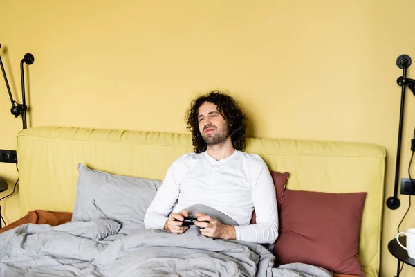 KYIV, UKRAINE - 25 AVRIL 2020 : jeune homme heureux tenant un joystick tout en jouant au jeu vidéo au lit — Photo de stock