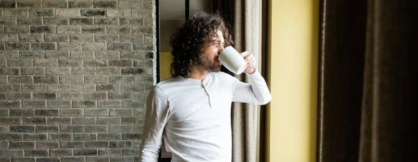 Horizontales Bild eines gutaussehenden Mannes im Pyjama, der Kaffee trinkt, während er morgens aus dem Fenster schaut — Stockfoto