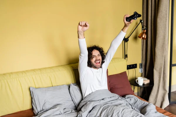 KYIV, UCRANIA - 25 DE ABRIL DE 2020: joven emocionado mostrando gesto ganador mientras juega videojuego en la cama - foto de stock