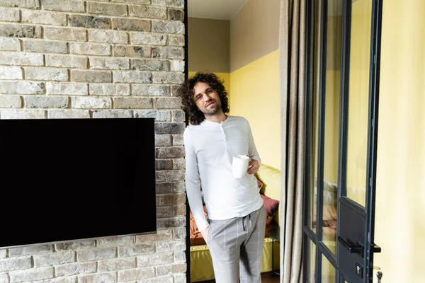 Handsome man in pajamas holding coffee cup and looking at camera near blank lcd screen hanging on brick wall — Stock Photo
