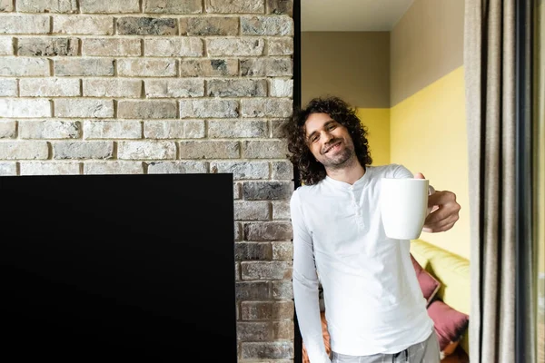 Hombre feliz sosteniendo la taza de café en la mano extendida cerca de pantalla lcd en blanco colgando en la pared de ladrillo - foto de stock