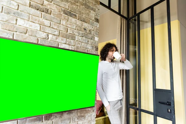 Giovane uomo in pigiama bere caffè mentre in piedi vicino schermo lcd verde appeso al muro di mattoni — Foto stock