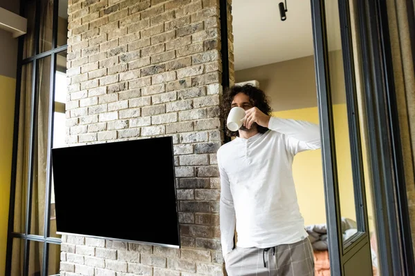 Junger Mann im Schlafanzug trinkt Kaffee, während er in der Nähe eines leeren LCD-Bildschirms steht, der an einer Ziegelwand hängt — Stockfoto