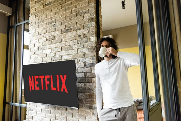 KIEW, UKRAINE - 25. April 2020: Junger Mann im Schlafanzug trinkt Kaffee, während er neben dem LCD-Bildschirm mit Netflix steht — Stockfoto