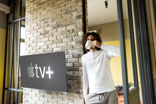 KYIV, UKRAINE - APRIL 25, 2020: young man in pajamas drinking coffee while standing near lcd screen with Apple TV — Stock Photo