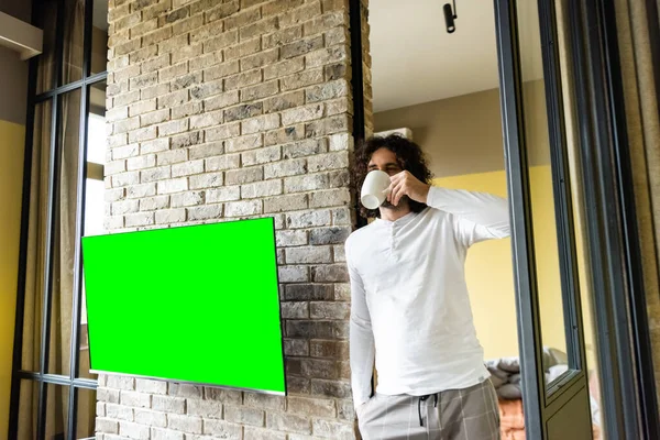 Junger Mann im Schlafanzug trinkt Kaffee, während er in der Nähe eines grünen LCD-Bildschirms steht, der an einer Ziegelwand hängt — Stockfoto
