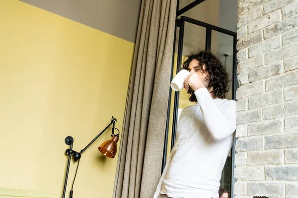 Молодой кудрявый мужчина в пижаме пьет кофе, стоя в спальне. — стоковое фото