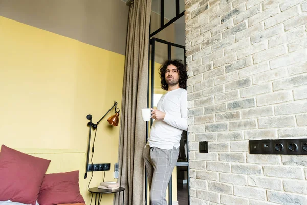 Handsome curly man in pajamas holding cup of coffee and looking away in bedroom — Stock Photo