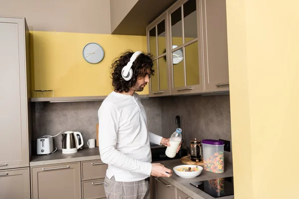 Joven en pijama y auriculares inalámbricos vertiendo leche en un tazón con hojuelas mientras prepara el desayuno - foto de stock