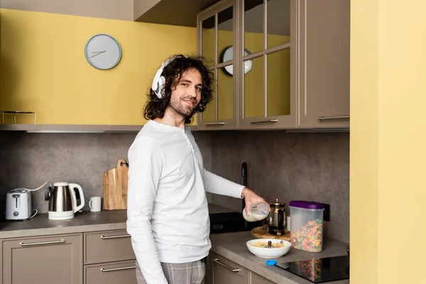 Uomo sorridente in cuffie senza fili versando latte in ciotola con fiocchi mentre guarda la fotocamera — Foto stock