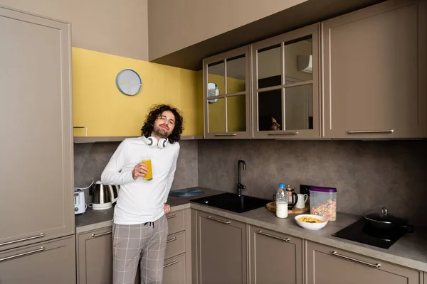 Homme souriant avec casque sans fil sur le cou boire du jus d'orange et regarder la caméra — Photo de stock