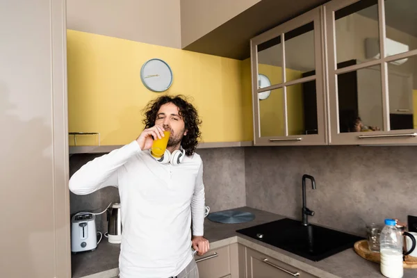 Schöner Mann mit drahtlosen Kopfhörern am Hals, Orangensaft trinkend und in die Kamera blickend — Stockfoto