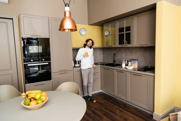 Verträumter junger Mann hört Musik in drahtlosen Kopfhörern mit geschlossenen Augen, während er Orangensaft in der Küche hält — Stockfoto