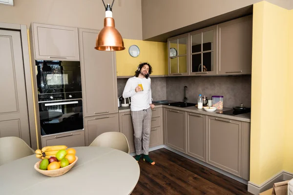 Dreamy young man listening music in wireless headphones with closed eyes while holding orange juice in kitchen — Stock Photo