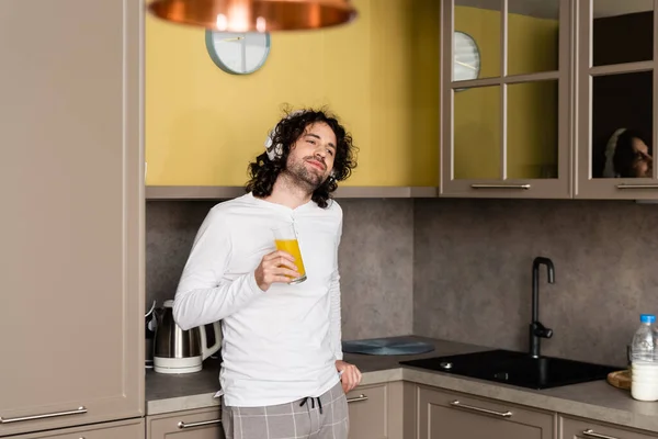 Homme souriant dans les écouteurs sans fil regardant loin tout en tenant du jus d'orange — Photo de stock