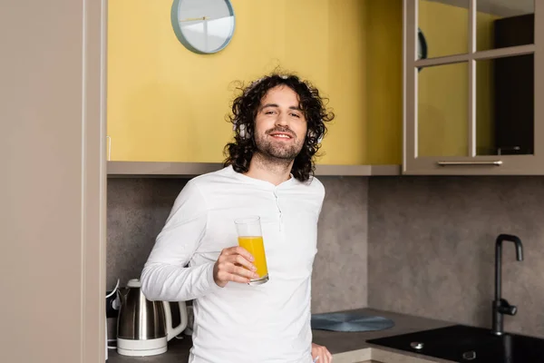 Hombre feliz en auriculares inalámbricos sosteniendo jugo de naranja mientras sonríe a la cámara - foto de stock