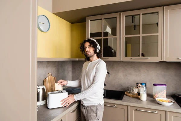 Lockiger Mann mit drahtlosen Kopfhörern, der Brot in den Toaster steckt, während er in der Küche in die Kamera schaut — Stockfoto