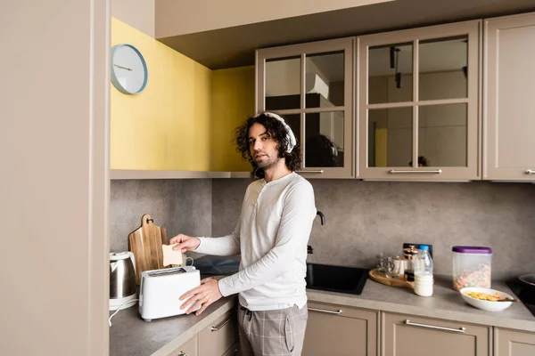 Bouclé homme dans les écouteurs sans fil mettre du pain dans grille-pain tout en regardant la caméra dans la cuisine — Photo de stock