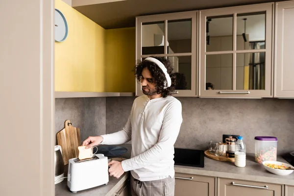 Hombre rizado en auriculares inalámbricos poner pan en la tostadora mientras se prepara el desayuno - foto de stock