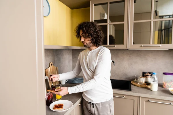 Frisé jeune homme en pyjama propagation confiture sur pain grillé pour le petit déjeuner — Photo de stock