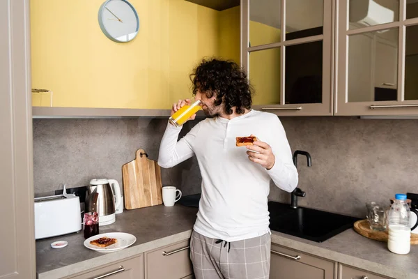 Joven en pijama bebiendo jugo de naranja y brindando con mermelada en la cocina - foto de stock