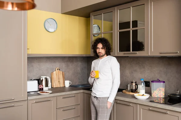 Uomo triste in pigiama guardando la fotocamera mentre tiene in mano un bicchiere di succo d'arancia in cucina — Foto stock