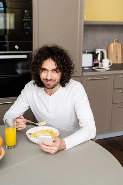 Hombre feliz sonriendo a la cámara mientras desayuna y chatea en el teléfono inteligente en la cocina - foto de stock
