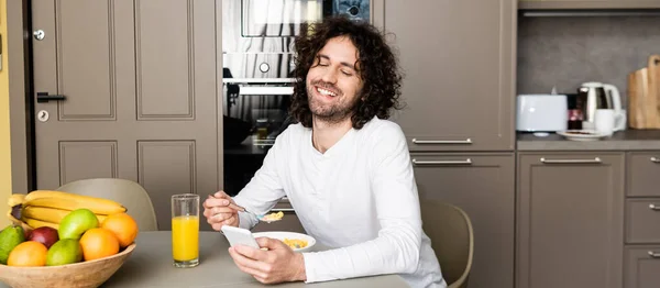 Colpo panoramico di uomo sorridente che chiacchiera sullo smartphone durante la colazione in cucina — Foto stock