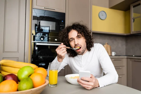 Handsome young man eating flakes and looking at camera while using smartphone during breakfast — Stock Photo