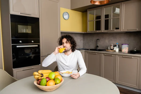 Красивый мужчина, смотрящий в камеру, пока пьет апельсиновый сок и пользуется смартфоном во время завтрака — стоковое фото