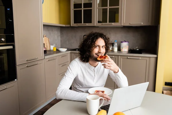 Cheerful freelancer eating toast and looking at camera near laptop and coffee cup — Stock Photo