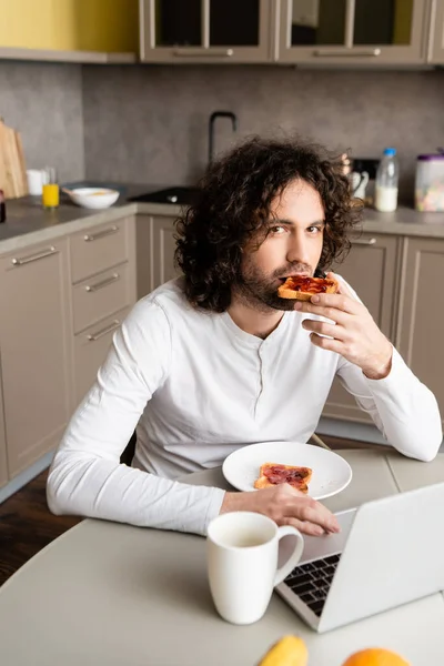 Handsome freelancer eating toast and looking at camera near laptop and coffee cup — Stock Photo