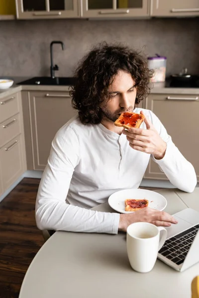 Freelancer pensativo comer torrada enquanto olha para laptop perto de xícara de café — Fotografia de Stock