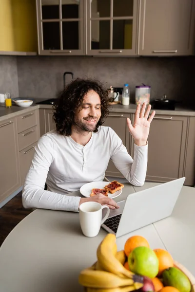 Feliz freelancer saludando la mano durante el chat de vídeo en el ordenador portátil cerca de la taza de café, tostadas y frutas frescas - foto de stock