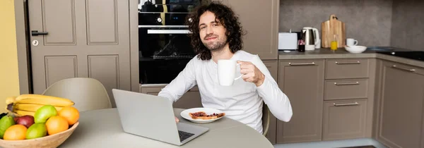 Immagine orizzontale di freelance sorridente che tiene la tazza di caffè e guarda la macchina fotografica vicino a computer portatile, pane tostato e frutta fresca — Foto stock