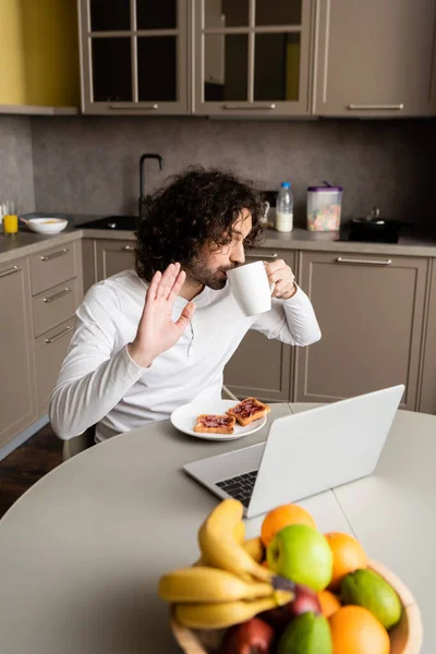 Freelance bouclé boire du café et agitant la main pendant le chat vidéo sur ordinateur portable dans la cuisine — Photo de stock