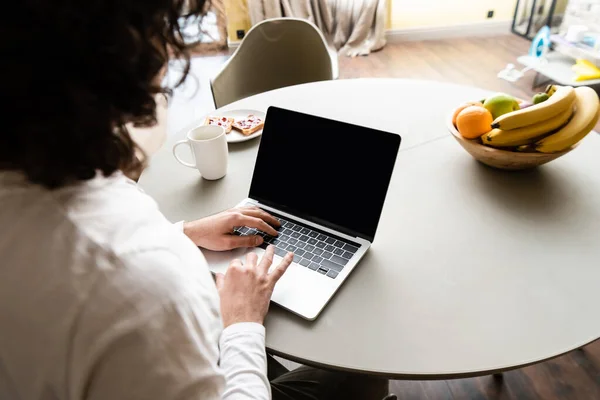 Back view of freelancer using laptop with blank screen near fruits, coffee cup and plate with toasts — Stock Photo