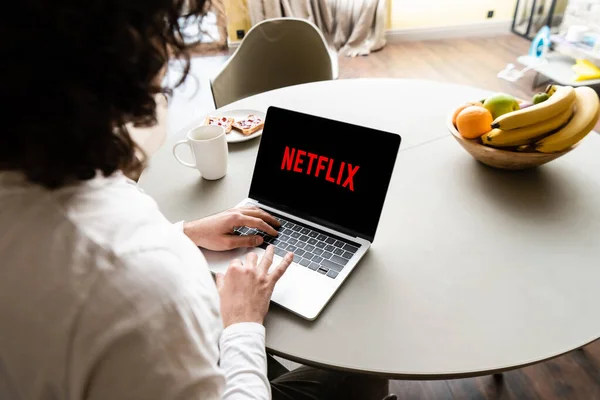 KYIV, UKRAINE - 25 AVRIL 2020 : vue arrière de pigiste à l'aide d'un ordinateur portable avec le site Web Netflix près de fruits, tasse de café et assiette avec des toasts — Photo de stock