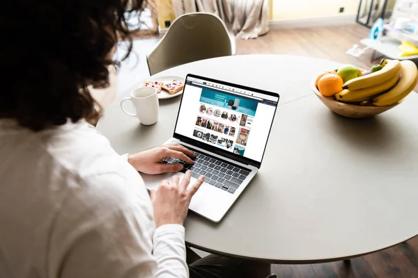 KYIV, UKRAINE - APRIL 25, 2020: back view of freelancer using laptop with Amazon website near fruits, coffee cup and plate with toasts — Stock Photo