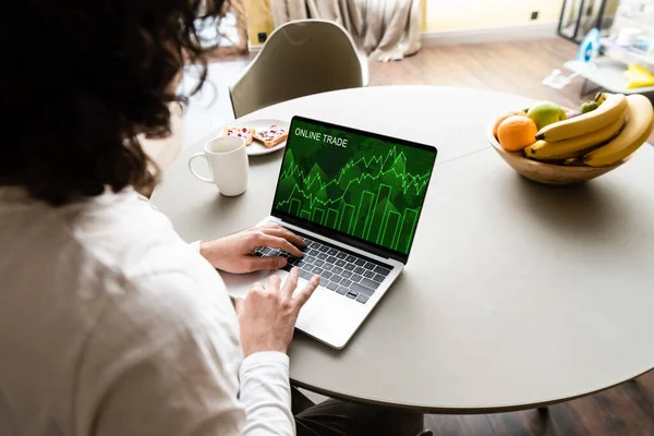 Back view of freelancer using laptop with online trade website near fruits, coffee cup and plate with toasts — Stock Photo