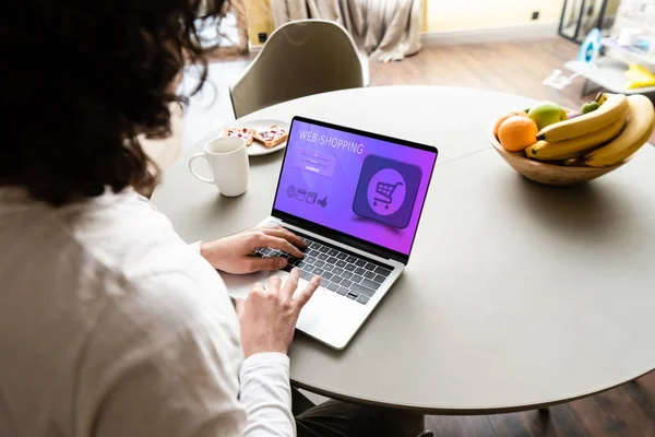 Back view of freelancer using laptop with web shopping website on screen near coffee cup, toasts and fresh fruits — Stock Photo