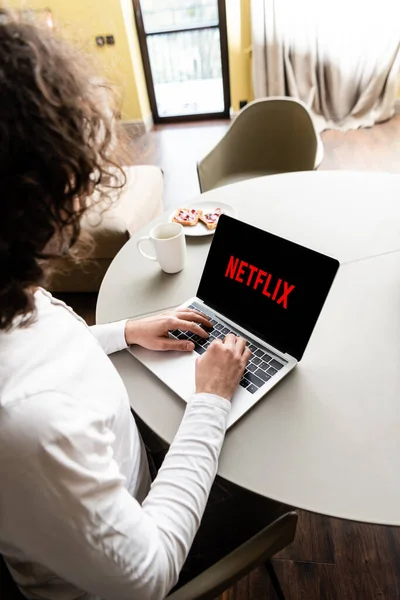 KYIV, UKRAINE - APRIL 25, 2020: high angle view of freelancer using laptop with Netflix on screen near coffee cup and plate with toasts — Stock Photo