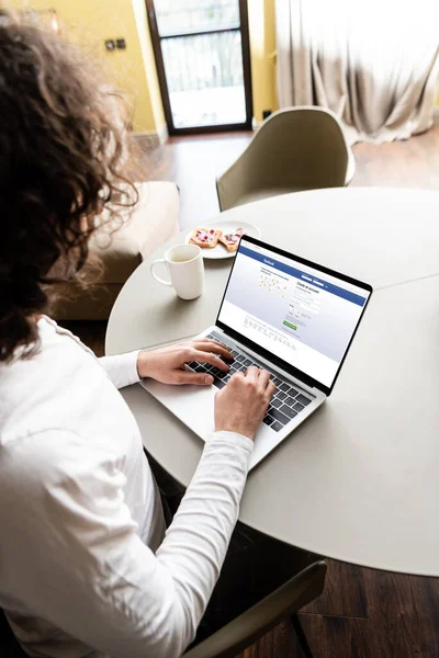 KYIV, UKRAINE - APRIL 25, 2020: high angle view of freelancer using laptop with Facebook on screen near coffee cup and plate with toasts — Stock Photo