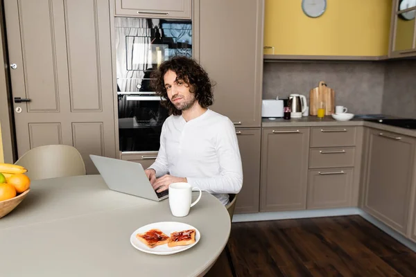 Jovem blogueiro olhando para a câmera enquanto digita no laptop perto da placa com saborosas torradas e xícara de café — Fotografia de Stock