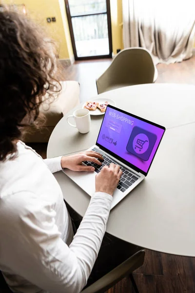 Visão de alto ângulo do freelancer usando laptop com site de compras na web perto de xícara de café e prato com torradas — Fotografia de Stock